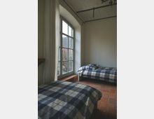 Ein einfaches Schlafzimmer mit zwei Einzelbetten, die mit karierten Bettbezügen ausgestattet sind. Der Raum hat eine große, bogenförmige Fensterfront mit weißen Vorhängen, die viel Tageslicht hereinlässt, und einen Boden aus Terrakottafliesen.