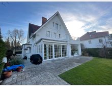 Zweistöckiges Einfamilienhaus mit weißer Fassade und dunklem Satteldach, großen Glasfenstern und Türen, umgeben von einem gepflasterten Terrassenbereich und einer gepflegten Rasenfläche. Im Hintergrund sind benachbarte Häuser und kahle Bäume sichtbar.