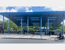 Bahnhof Neuenhagen bei Berlin mit moderner, geschwungener Überdachung aus blauen Stahlträgern und Gitterpanels. Im Vordergrund ein gepflasterter Gehweg, wenige Passanten sowie ein Radfahrer, im Hintergrund Grünflächen.