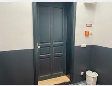 Dunkelgraue Tür in einem Flur mit weiß-grauen Wänden; an der Wand ist ein orangefarbener Brandmelder sowie ein Hinweisschild mit der Aufschrift „Rauchabzug“ angebracht. Im unteren rechten Bereich steht ein weißer Abfalleimer.