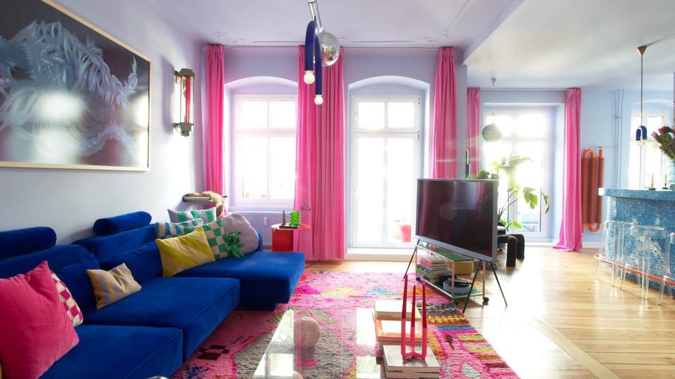 Bright, modern living room with a blue sectional sofa and colorful pillows, a glass coffee table on a patterned pink rug, and a TV on a stand. Large white-framed windows and a glass door are framed by pink curtains, with pendant lights and an open area leading toward a kitchen or dining space.