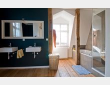 Modernes Badezimmer mit einer dunklen Wand, an der zwei weiße Waschbecken mit rechteckigem Spiegel hängen. Rechts befinden sich eine Badewanne, eine Glasdusche und ein Fenster, das Tageslicht hereinlässt, kombiniert mit sichtbaren Holzbalken und einem Holzboden.