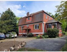 Ein zweistöckiges Haus in Rot mit roten Dachziegeln steht in einer ländlichen Umgebung. Im Vordergrund befinden sich ein unbefestigter Weg, ein kleiner bepflanzter Bereich und ein Hof mit geparkten Autos, umgeben von Bäumen und Sträuchern.