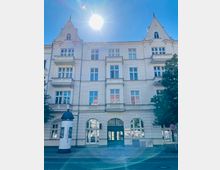 Mehrstöckiges helles Backsteingebäude mit symmetrischer Fassade, hohen Fenstern, kleinen Balkonen und zwei spitzen Giebeln; im Erdgeschoss befindet sich ein zentraler Eingangsbereich mit Bogenfenster. Davor verläuft eine Straße mit einem Litfaßsäulen-Plakat und Bäumen am rechten Rand unter blauem Himmel mit Sonne.