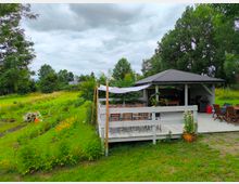 Ein Garten mit einer offenen Terrasse, die mit einem Pavillon und Sitzgelegenheiten ausgestattet ist. Im Vordergrund befinden sich Beete mit Blumen und Gemüse, umgeben von Rasen und Bäumen im Hintergrund.