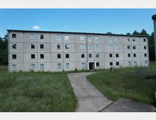 Ein vierstöckiges, graues Plattenbaugebäude mit zahlreichen quadratischen Fenstern in einer ländlichen, grasbewachsenen Umgebung. Ein betonierter Weg führt von vorne zur Eingangstür des Gebäudes. Im Hintergrund sind Bäume zu sehen.