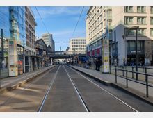 Straßenbahnhaltestelle vor dem Bahnhof Friedrichstraße in Berlin, umgeben von modernen Gebäuden aus Glas und Stein. Im Hintergrund sind Bahngleise, eine Fußgängerbrücke und Schilder für S- und U-Bahn erkennbar.