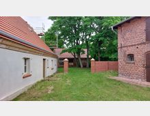 Hof mit Gebäuden aus rotem Ziegelstein und verputzten Fassaden, flankiert von einem Rasen und umgeben von Bäumen; links ein langes Gebäude mit roten Dachziegeln, rechts ein Ziegelbau mit Fenstergittern. Im Hintergrund sind ein weiteres kleines Gebäude und ein Zaun zu sehen.