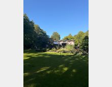 Großer Garten mit weitläufiger Rasenfläche, Blumen- und Strauchbeeten sowie hohen Bäumen am Rand. Im Hintergrund steht ein flaches, modernes Gebäude mit markant gewelltem Dach und einer Terrasse mit Sonnenschirmen.