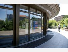 Eine moderne Glasfassade mit leicht gebogenem Dach und großen Fensterscheiben, die Plakate und Informationen zeigen. Im Hintergrund befinden sich ein Park mit Bäumen sowie historische und moderne Gebäude.