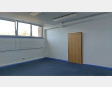 Leerer Raum mit blauem Teppichboden, weiß gestrichenen Wänden und einer Holztür auf der rechten Seite. Oben befinden sich rechteckige Fenster, die Tageslicht hineinlassen, sowie ein weißer Heizkörper an der Wand.