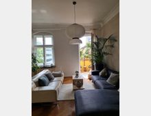 Wohnzimmer mit hellen Wänden und einem Holzboden, ausgestattet mit einer weißen und einer dunkelgrauen Couch, einem rechteckigen Couchtisch sowie einer großen Papier-Hängeleuchte. Im Hintergrund befinden sich hohe Fenster und eine Tür zu einem kleinen Balkon mit Pflanzen.