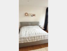 Ein Schlafzimmer mit einem grauen Polsterbett, bedeckt von einer Bettdecke mit Rautenmuster. Über dem Bett hängt ein Wandregal mit dekorativen Objekten und Bildern, rechts ist ein Fenster mit dunklen Vorhängen zu sehen, der Boden besteht aus Holz.