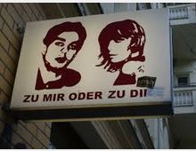 Leuchtwerbeschild mit der Aufschrift "Zu mir oder zu dir" zeigt stilisierte Porträts eines Mannes und einer Frau in Rot auf weißem Hintergrund; es ist an einer Gebäudewand angebracht. Im Hintergrund sind Balkone und Gebäudefassaden erkennbar.