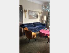 Wohnzimmer mit einer blauen Samtcouch, einem runden Tisch mit rotem Tischtuch und zwei gelben Polsterstühlen. An der Wand hängen goldene Wandleuchten und ein Landschaftsbild, ergänzt durch eine Stehlampe und einen Kronleuchter an der Decke.