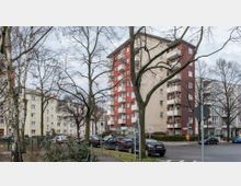 Straßenszene mit mehrstöckigen Wohngebäuden, darunter ein rot-beiges Gebäude mit Balkonen, umgeben von kahlen Bäumen im Winter. Im Vordergrund sind parkende Autos, Gehwege und Straßenschilder zu sehen.