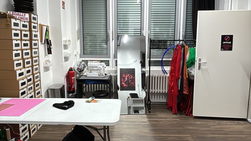 Small studio workspace with a wood floor, a folding table, storage drawers, and sewing equipment, facing a wall of tall windows with shutters. Exposed ceiling pipes and a fluorescent light hang overhead, with costume racks and a white door along the right wall.