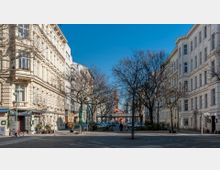 Städtischer Platz mit gepflastertem Boden und kahlen Bäumen, umgeben von mehrstöckigen historischen Wohngebäuden mit heller Fassade. Im Hintergrund ist eine rote Backsteinkirche mit einem spitzen Turm sichtbar.