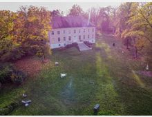 Luftaufnahme eines hellrosa Herrenhauses mit weißen Fensterrahmen, umgeben von einem weitläufigen, grünen Park mit herbstlich gefärbten Bäumen. Im Vordergrund befinden sich vereinzelte Gartenmöbel auf der Wiese, die mit Laub bedeckt ist.