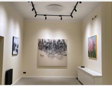 Ausstellungsraum mit beigefarbenen Wänden und moderner Beleuchtung. Drei Gemälde hängen an den Wänden, darunter eine großformatige, abstrakte Arbeit in der Mitte; an der rechten Seite steht eine weiße Kommode.