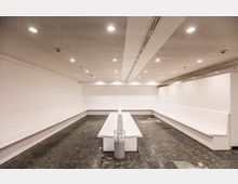 Minimalistischer, weißer Innenraum mit langen, an den Wänden befestigten Sitzbänken und einem zentralen Tisch mit einem kleinen Mülleimer darauf; der Raum hat einen Boden aus poliertem dunklem Stein und eine Decke mit eingelassenen Deckenlampen.