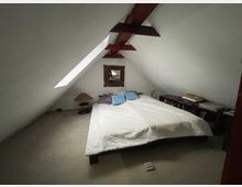 Schlafzimmer im Dachgeschoss mit schrägen weißen Wänden und sichtbaren Holzbalken, in der Mitte ein Doppelbett auf Teppichboden. An den Seiten stehen niedrige Möbel und Gegenstände, an der Stirnwand ein kleines Fenster.