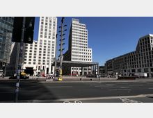 Der Potsdamer Platz in Berlin mit modernen Hochhausfassaden und dem gläsernen Eingang des Bahnhofs "Potsdamer Platz" im Zentrum. Im Vordergrund sind Straßen und Gehwege erkennbar, während Passanten und Fahrzeuge die Szene beleben.