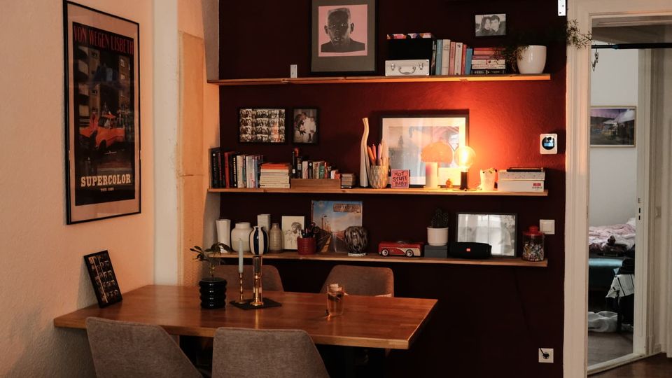 Gemütlicher Essbereich in einer Wohnung mit Holztisch und Polsterstühlen vor einer dunkelroten Wand mit drei langen Wandregalen voller Bücher, Bilderrahmen, Vasen und einer kleinen Lampe. Links hängt ein gerahmtes Poster, rechts führt eine Türöffnung in ein angrenzendes Zimmer.