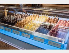 Ein beleuchteter Verkaufstresen mit einer Auswahl an Eis am Stiel in verschiedenen Farben und Geschmacksrichtungen, wie Schokolade, Vanille, Honig-Lavendel, und Joghurt-Heidelbeere, jeweils in getrennten Fächern präsentiert. Im Hintergrund ist ein Verkäufer sichtbar, während die Geschmacksrichtungen auf Schildern am unteren Rand des Tresens angegeben sind.