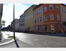 Straßenszene mit mehrstöckigen Wohn- und Geschäftshäusern in verschiedenen Farben, darunter Orange und Gelb, entlang einer gepflasterten Straße. Im Vordergrund sind Straßenschilder, ein Gehweg und ein Laternenmast sichtbar.