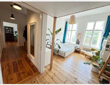 Helles Schlafzimmer mit Holzfußboden, großen weißen Fenstern und blauen Vorhängen, eingerichtet mit einem Bett, Sessel, Pflanzen und kleinen Möbelstücken. Im Vordergrund ein Übergang zu einem Flur mit weiteren Holzböden und dezenten Hängelampen.