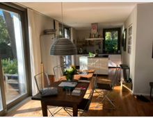 Helles Ess- und Küchenzimmer in einem modernen Wohnhaus mit großen Glasfenstern, die auf einen Garten blicken. Der Raum zeigt einen Holz-Esstisch mit Stühlen, eine hängende Lampe, eine offene Küche mit weißen Schränken und eine parkettierte Holzbodenfläche.