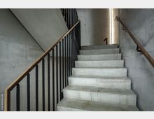 Betontreppe in einem modernen Treppenhaus mit minimalistischer Gestaltung, ergänzt durch schwarze Geländer mit Holzhandläufen. Die Wände sind aus Sichtbeton, und ein schmales Fensterband lässt Licht in den Raum.