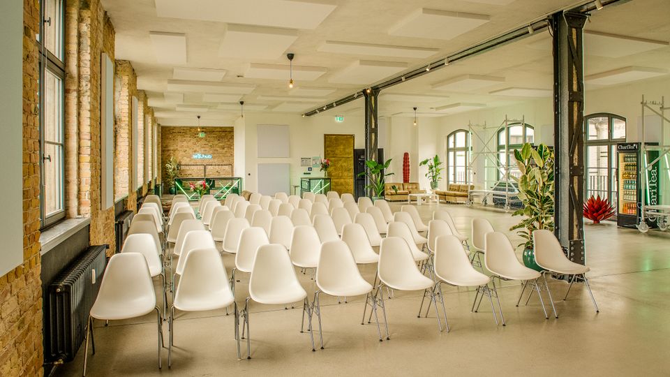 Großer, heller Veranstaltungsraum im Industrie-Loft-Stil mit Reihen weißer Stühle, hohen Fenstern und freiliegenden Backsteinwänden. Sichtbare Stahlträger, Akustikdeckenpaneele, Pflanzen sowie Sitzbereiche und Getränkeautomaten im Hintergrund.