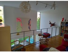 Ein modernes Wohnzimmer mit heller Holzbodenfläche und einer Galerieebene, die durch ein Metallgeländer abgegrenzt ist. An der weißen Wand hängen eine bunte Pop-Art-Malerei und eine stilisierte Hirschkopf-Dekoration; daneben steht ein Bücherregal und ein Servierwagen. Große Fenster lassen natürliches Licht herein und bieten Ausblick auf einen Garten.