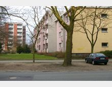 Wohngebiet mit mehrstöckigen Wohnhäusern in hellen Farben, umgeben von einer Grünfläche mit Bäumen. Im Vordergrund befindet sich ein einzelnes geparktes Auto auf einem unbefestigten Bereich neben der Straße.