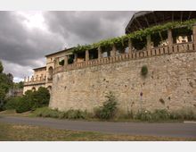 Historische Steinmauer mit hochliegender Arkadengalerie, die von Reben bewachsen ist, im Vordergrund eine Straße und wild bewachsene Vegetation; im Hintergrund ein Gebäude mit großen Fenstern unter bewölktem Himmel.