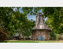 Eine historische Windmühle aus Backstein mit dunkler Holzverkleidung und Flügeln, umgeben von Bäumen in einer grünen Parklandschaft. Vor der Mühle befindet sich ein Eingang mit der Aufschrift „Vereinsheim“ und dekorativen Elementen.