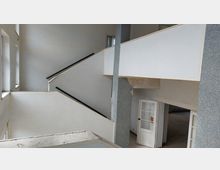 Ein Treppenhaus mit weißen Wänden und schwarzem Handlauf in einem scheinbar verlassenen Gebäude. Links befinden sich große Fenster, rechts unten eine weiße Tür mit Glasprofilen, und einige Stellen an den Wänden sind unvollständig renoviert.