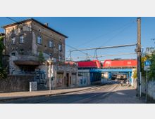 Eine Straßenansicht mit einer alten, verfallenen Gebäudefassade links, Graffiti darauf, darunter Fußweg und Straßenbahnschienen. Im Hintergrund fährt ein roter Zug der Deutschen Bahn über eine blaue Eisenbahnbrücke.