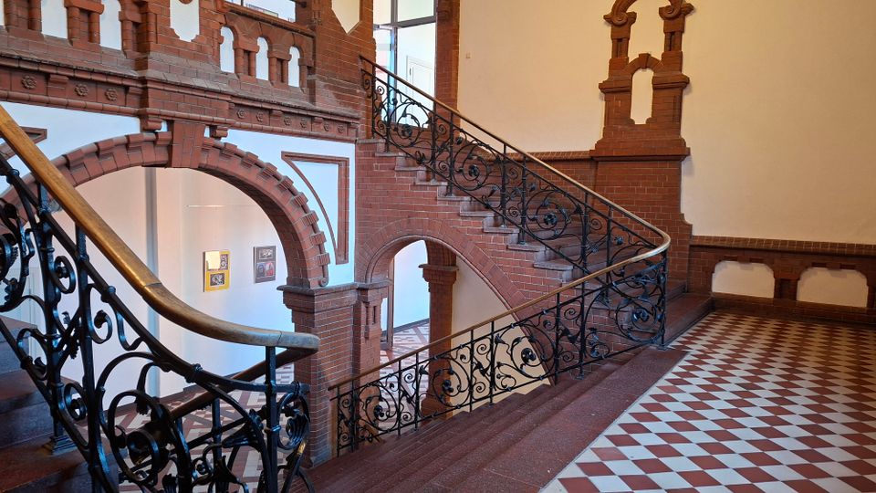 Innenansicht eines Treppenhauses mit roten Ziegelbögen und dekorativem Mauerwerk, einer geschwungenen Treppe und einem schwarzen schmiedeeisernen Geländer. Der Boden ist mit rot-weißen Schachbrettfliesen ausgelegt, an den Wänden hängen gerahmte Bilder.