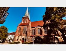 Backsteinkirche mit hohen Spitzbogenfenstern und einem Turm mit spitzen Doppeltürmen, umgeben von Bäumen und angrenzenden Gebäuden vor einem klaren, blauen Himmel.