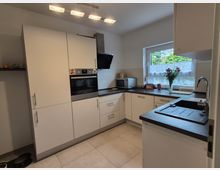 Moderne, helle Einbauküche in L-Form mit weißen Schränken, integriertem Backofen, schwarzer Arbeitsplatte und Edelstahlspüle. Großes Fenster über der Arbeitsfläche mit Blick ins Grüne sowie Deckenleuchte und Fliesenboden.
