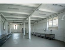 Weiß gefliester, leerer Raum mit Industrieküchen-Ausstattung, einschließlich Edelstahl-Arbeitstischen, Spülbecken und freiliegender Deckenbeleuchtung. Große Fenster lassen Tageslicht herein; sichtbare Verrohrung und Wände aus Beton tragen zur industriellen Atmosphäre bei.