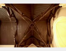 Holzdecke einer Kirche mit dunklen Holzbalken und Spitzbogenkonstruktionen, die diagonal verlaufen. Die Konstruktion wird von filigranen Metallverstrebungen ergänzt und rechts ist ein Teil einer hell beleuchteten Wand sichtbar.