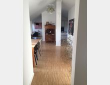 Wohnraum mit modernem Design, hellem Holzfußboden und weißen Wänden. Im Hintergrund stehen ein antiker Schreibtisch mit Kerzenständer und ein eingebauter Küchenschrank, während im Vordergrund ein Esstisch mit Stühlen und eine Zimmerpflanze sichtbar sind.