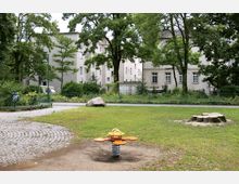Ein kleiner Spielplatz in einem begrünten Stadtpark mit einem Wipptier in der Mitte, umgeben von Rasen, vereinzelten Bäumen und gepflasterten Wegen. Im Hintergrund sind mehrstöckige Wohnhäuser und dichte Vegetation zu sehen.