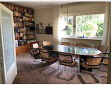 Wohnzimmer oder Besprechungsraum mit einem ovalen Glastisch und acht Stühlen mit geflochtenen Sitzflächen. Im Hintergrund befindet sich ein großes Regal voller Bücher, ein breites Fenster gibt den Blick auf grüne Natur frei, und darunter ist ein Heizkörper erkennbar.