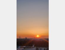 Städtische Skyline bei Sonnenuntergang mit einem klaren Horizont und orangefarbenem Himmel; mehrere Dächer und Schornsteine im Vordergrund, sowie Kirchtürme und Gebäude am Horizont.