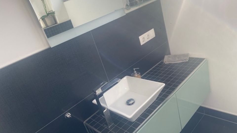 Modernes Badezimmer-Waschbecken auf einer dunkel gefliesten Ablage mit mintfarbenen Unterschränken und schwarzer Wandverkleidung. Darüber befindet sich ein Fenster mit kleiner Ablagefläche, auf der verschiedene Pflegeartikel stehen.